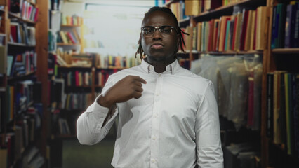 Man young african american in white button shirt points to chest and tugs collar amid crowded bookshelves in building  confidence focus. © Krakenimages.com