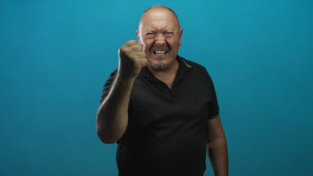Senior hispanic man clenches fist, bares teeth and raises forearm in studio with blue backdrop; anger defiance resilience.