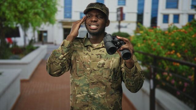 Young soldier man in camouflage holding binoculars salutes with right hand to forehead on a building walkway with railings and planters; duty vigilance.