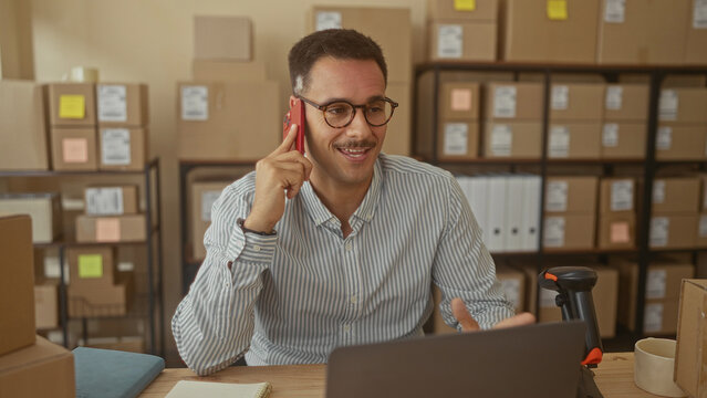 Man holding smartphone to ear in building stockroom with laptop, barcode scanner and stacked cardboard boxes on shelves; entrepreneurial focus.