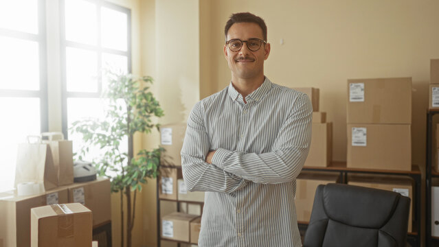 Man with arms crossed beside stacked shipping boxes in a building, wearing striped shirt and glasses; small business confidence.