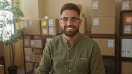 Man with beard and glasses smiles amid stacked cardboard boxes and shelves in a building office; entrepreneurial confidence.