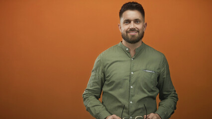 Obraz premium Hispanic man holding eyeglasses with hands in studio orange backdrop, raising frames to face and smiling while adjusting glasses; approachable.