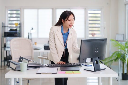 Asian adult businesswoman professional working hard standing at modern office desk using desktop computer diligent employee concentrating on work tasks