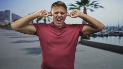 Naklejka premium Man plugging fingers in ears on a marina promenade wearing a red polo shirt, grimacing and leaning slightly forward while blocking loud harbor noise; annoyance.