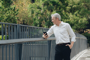 Fototapeta premium Mature man in white shirt standing outdoors by railing holding smartphone, looking away thoughtfully in green park setting on sunny day