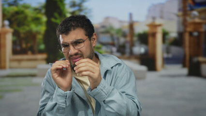 Young hispanic man in denim jacket and glasses shields face with hands while adjusting frames on busy city street; fear tension.