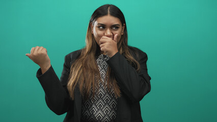 Woman covers mouth with hand while gesturing thumb back toward unseen object and stands before a green studio backdrop  playful surprise. © Krakenimages.com