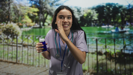 Fototapeta premium Woman nurse in lavender scrubs wearing stethoscope and badge holding blue pill bottle with hand to cheek and smiling in park; reassurance care.