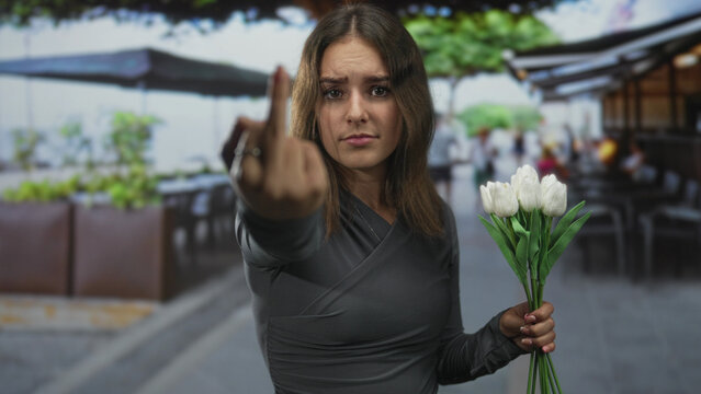 Young hispanic woman showing middle finger to camera while holding bouquet of white tulips on a restaurant terrace by the street; defiance.