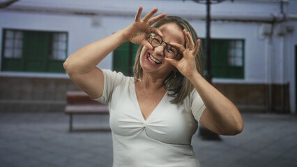 Fototapeta premium Woman frames eyes with hands while smiling and wearing glasses in a courtyard with bench and building facade; playful joy.