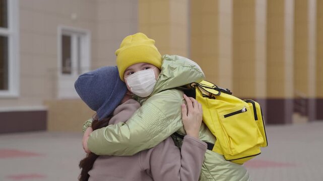 child hugs mother, mask, mother escorts her daughter school lesson, funny learning activities for pleasure, little kid with school backpack near school building, happy family, children s learning time