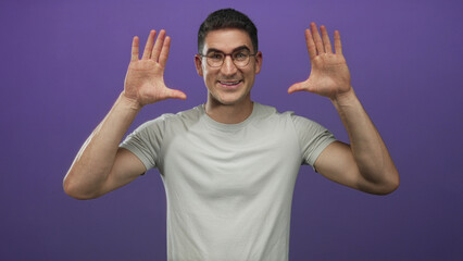 Fototapeta premium Man raising both hands with palms facing forward wearing glasses and light gray t shirt in purple studio backdrop; playful confidence.