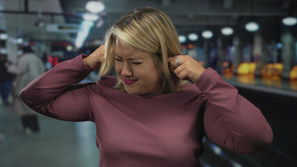 Fototapeta premium Young blonde woman pressing her hands on her ears at a noisy crowded train station platform; anxiety.
