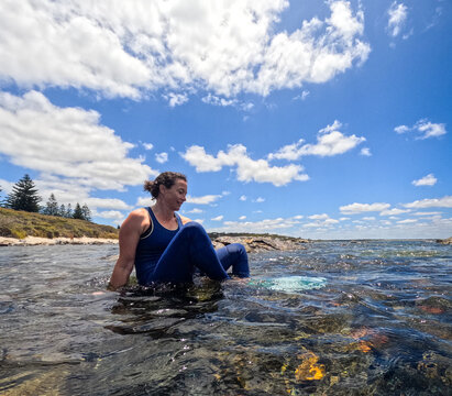 Woman in wetsuit wearing mask, snorkel and fins sitting in clear ocean water preparing to snorkel