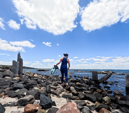 Woman in blue wetsuit walking along rocky beach coastline holding snorkelling fins & mask