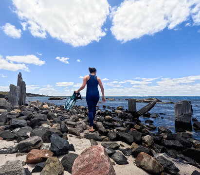 Woman in blue wetsuit walking along rocky beach coastline holding snorkelling fins & mask