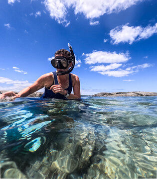 Woman in wetsuit wearing mask, snorkel and fins sitting in clear ocean water preparing to snorkel