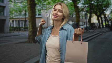 Fototapeta premium Woman holding a pink shopping bag, thumb pointing to chest, eyes closed and smiling on a city street; confidence.