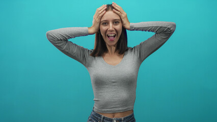 Young woman with hands on head and open mouth, wearing gray long sleeve top and jeans with visible...
