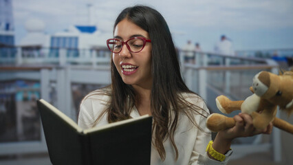 Hispanic woman on a boat reads a book while holding a stuffed toy, enjoying a seaside view...