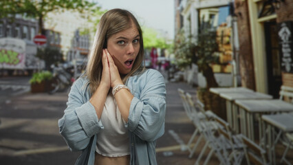 Fototapeta premium Woman pressing hands to cheek gesture on street terrace with urban cafe seating and blurred building facade; surprise.