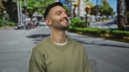 Fototapeta premium Young hispanic man smiling on an urban street with trees and buildings in the background, embodying a relaxed and content vibe on a sunny day outdoors.