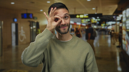 Fototapeta premium Hispanic man making ok gesture in airport terminal, exuding confidence and style with casual olive sweater amid lively travel ambiance.