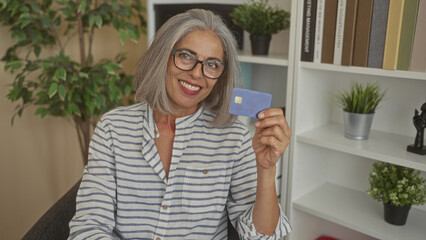 Woman holding a blue creditcard and smiling in an office building with shelves and plants;...