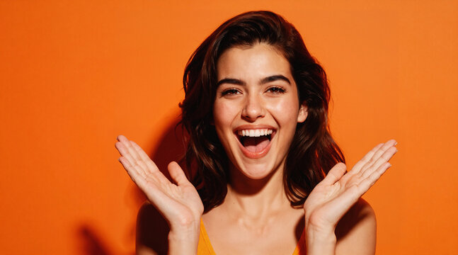 A woman with a cheerful expression ofA smiling woman with a cheerful expression, laughing with open mouth wide holding hands in front of her face,  in front of an orange background