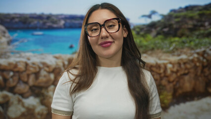Young hispanic woman wearing glasses and white shirt hand on chest on street with seaside wall;...