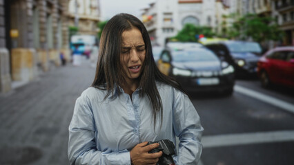 Obraz premium Hispanic woman clutching a camera at a crosswalk on a busy city street, wincing with a pained grimace while cars wait behind her; distress.