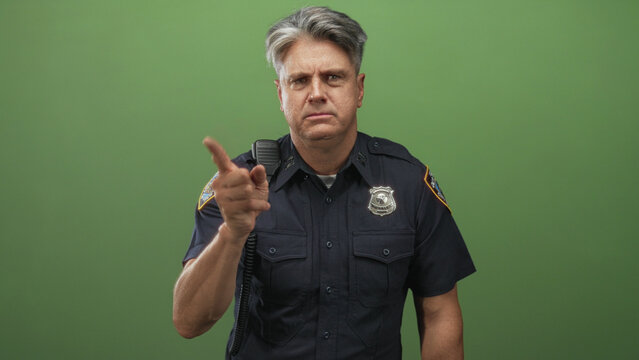 Man police officer points finger at camera in studio greenscreen, badge and shoulder radio visible; authority command.