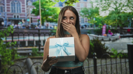 Fototapeta premium Young woman holding a gift box, hand to mouth beside canal in front of building with boat and trees; surprise celebration.