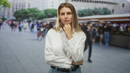 Fototapeta premium Woman with hand on chin and folded arm wearing white shirt in a crowded street market near stalls and buildings; quiet contemplation.