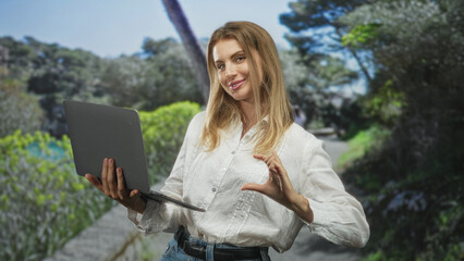 Blonde woman holding laptop and making heart hand gesture while standing on a sunlit forest path wearing white shirt; playful confidence.