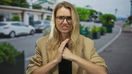 Fototapeta premium Woman displaying gesture angrily on city street with cars in background blonde caucasian wearing glasses and beige blazer during daytime