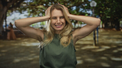 Woman expressing stress outdoors in a sunny park setting surrounded by trees, wearing casual green...