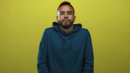 Fototapeta premium Man face shrugs shoulders in yellow studio wearing blue hoodie, beard visible, eyes closed and shoulders raised in a tense shrug; uncertainty.