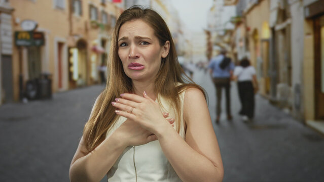 Woman clutch chest in street with agonized expression and tearful gaze amid blurred city buildings and distant passersby; pain.