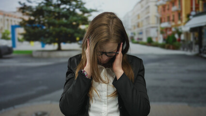 Fototapeta premium Young woman wearing glasses and blazer holds her ears in a busy city street with blurred storefront buildings; distress.