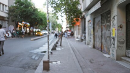 Blurred urban street scene in shallow bokeh showing sidewalk, graffiti covered storefronts, trees and distant lights in soft defocused background; background backplate copyspace calm.