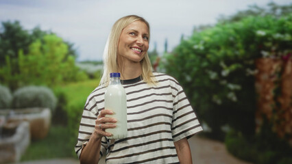 Fototapeta premium Woman holding milk bottle with hand, smiling while posing in studio garden set; contentment wholesome health.