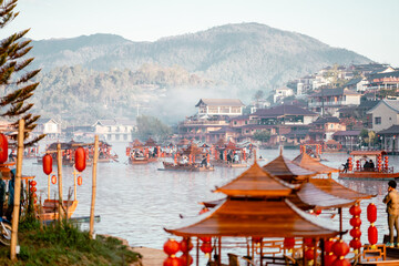 Obraz premium Crowded Wooden Boats with Red Lanterns on Misty Morning Lake with Chinese Village and Mountain Backdrop, Ban Rak Thai