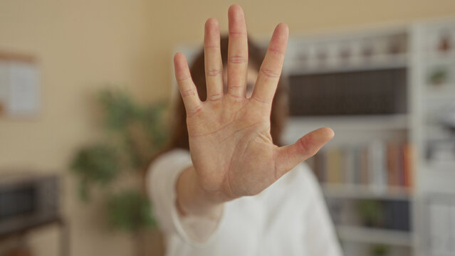 Woman raising outstretched hand, palm to camera in office wearing glasses and white blouse near bookshelf; personal boundary defiance.