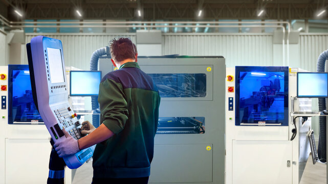 Manufacturing plant. Technician operates CNC machine via control panel, monitoring automated production process on industrial equipment in modern factory workshop.