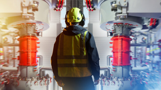 Industrial plant supervision, process equipment monitoring. Technician in safety helmet and vest inspects high pressure machinery and pipeline systems inside an energy facility.