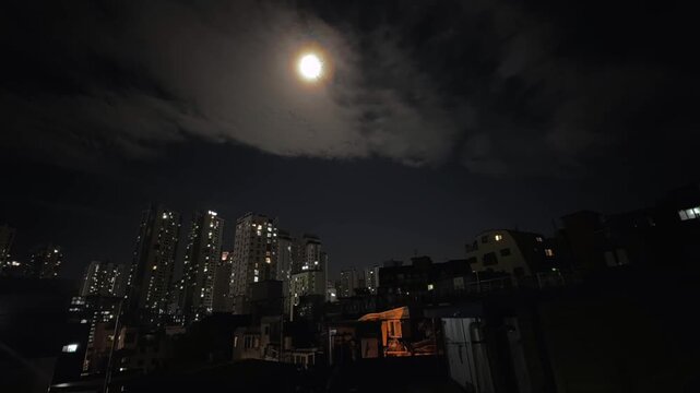 Full moon above residential city skyline with fast moving clouds across night sky, illuminated apartment buildings under dramatic moonlight creating atmospheric urban night scene.