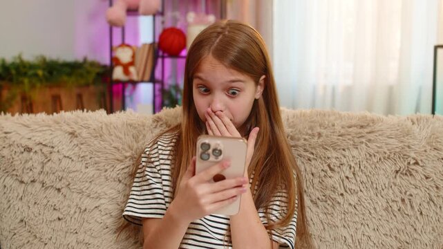 Young child girl sits on sofa reads surprising message on smartphone, reacts with wide open eyes curious expression. School kid looks stunned by unexpected content holding device tightly in both hands