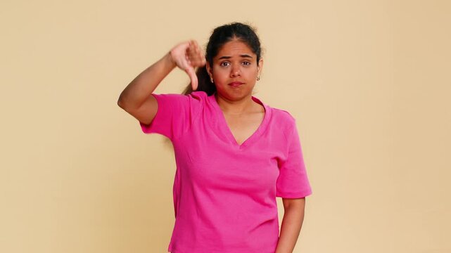 Indian woman lightly shakes head with clear disappointment, lowers shoulders shows thumbs down. Girl on beige background clearly indicates her disagreement failure or unhappiness demonstrating dislike